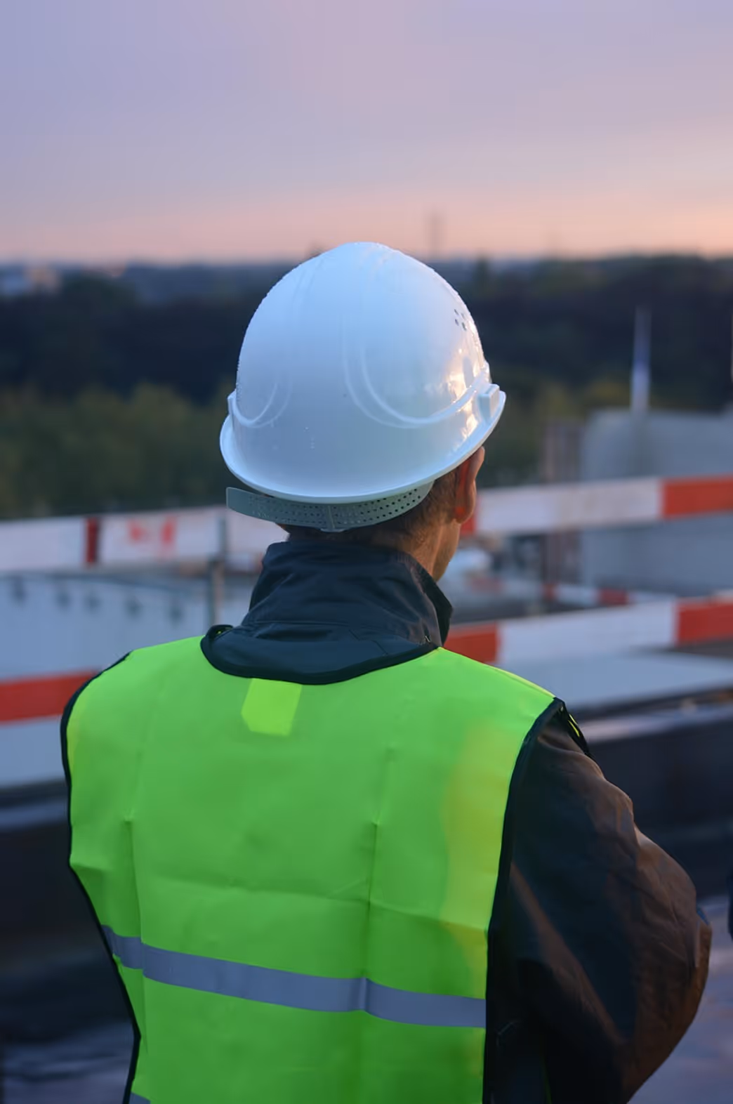 Construction worker wearing a white hard hat and neon safety vest overlooking a site at dusk.