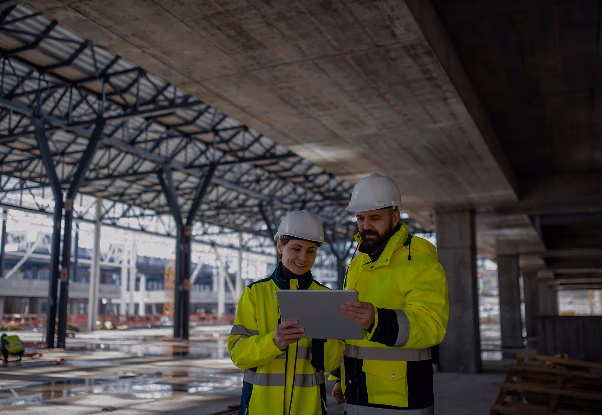 Two construction workers in yellow jackets and helmets looking at a tablet inside a building site.