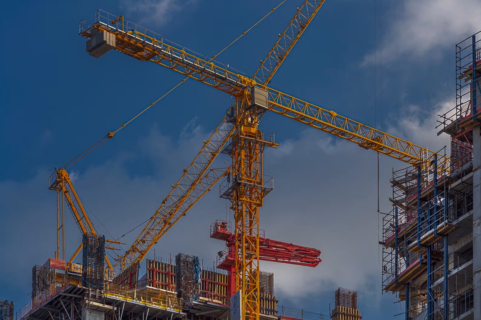 Yellow construction cranes over a building under construction with scaffolding and a cloudy blue sky.