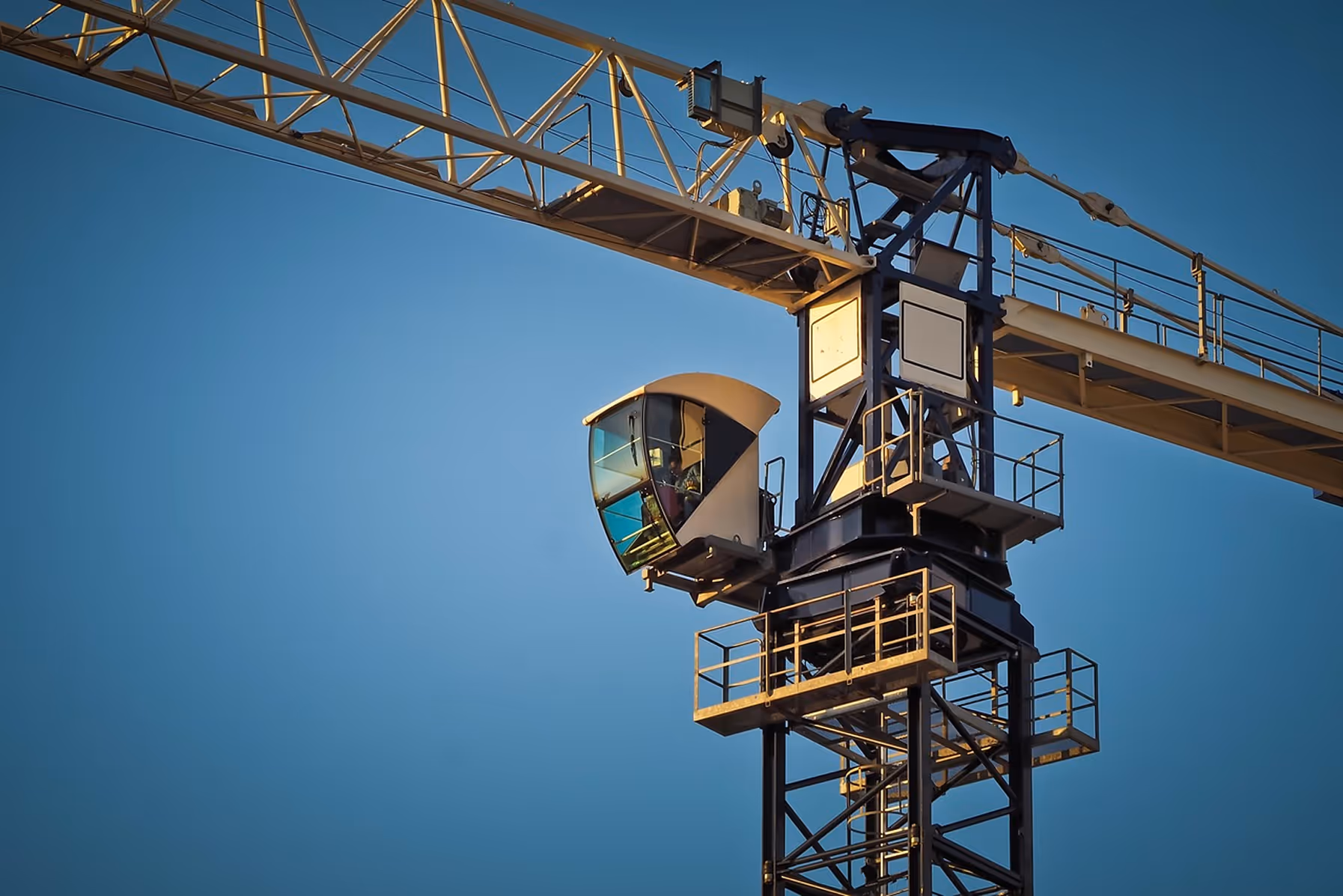 Crane operator cabin on a tall construction crane against a clear blue sky.
