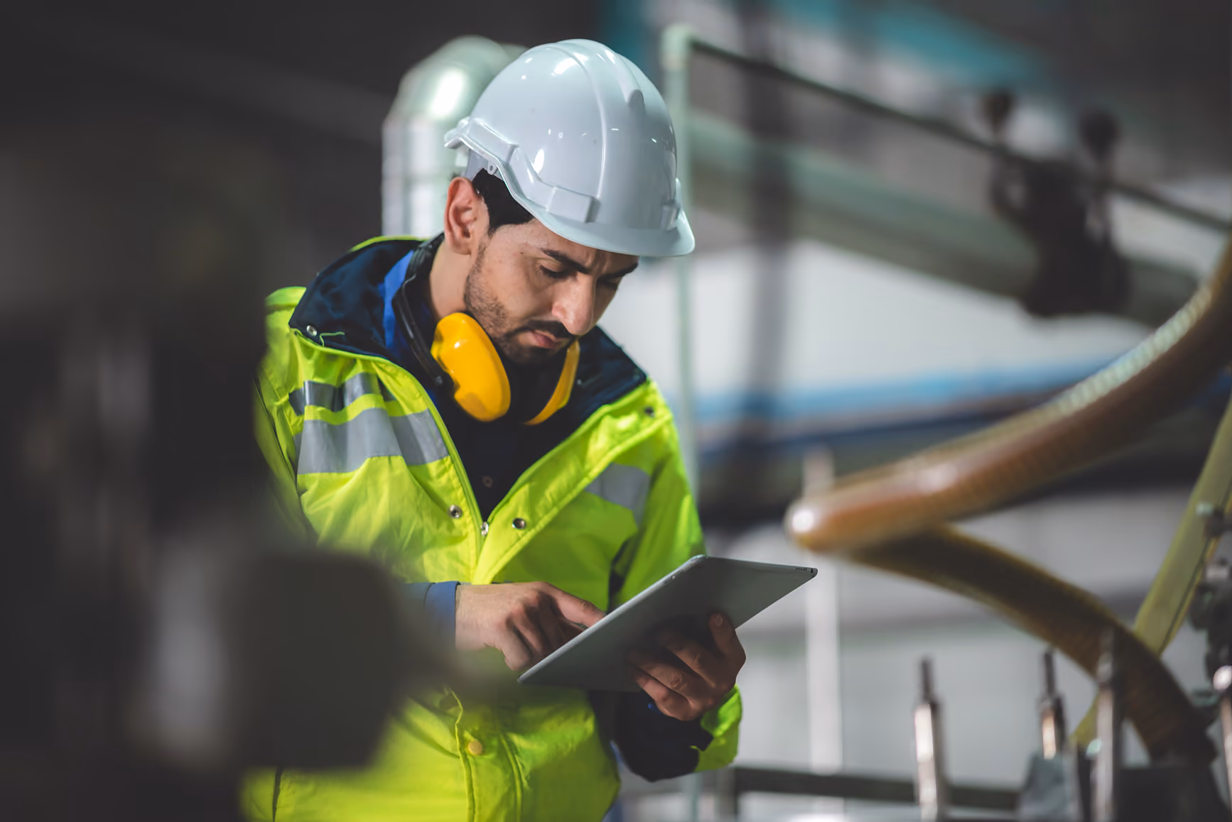 Male worker in helmet and high-visibility jacket using tablet device in industrial setting.
