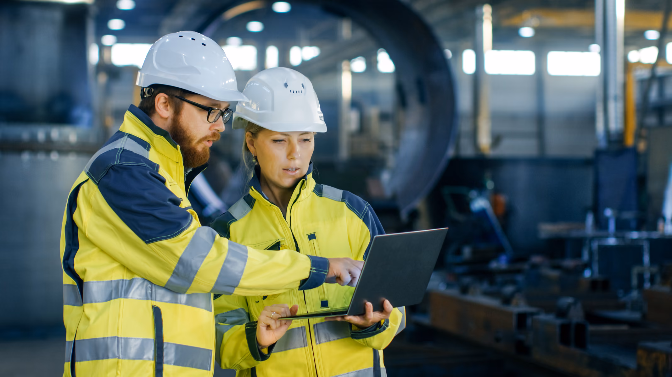 Two engineers wearing hard hats and high-visibility jackets looking at a laptop in an industrial setting.