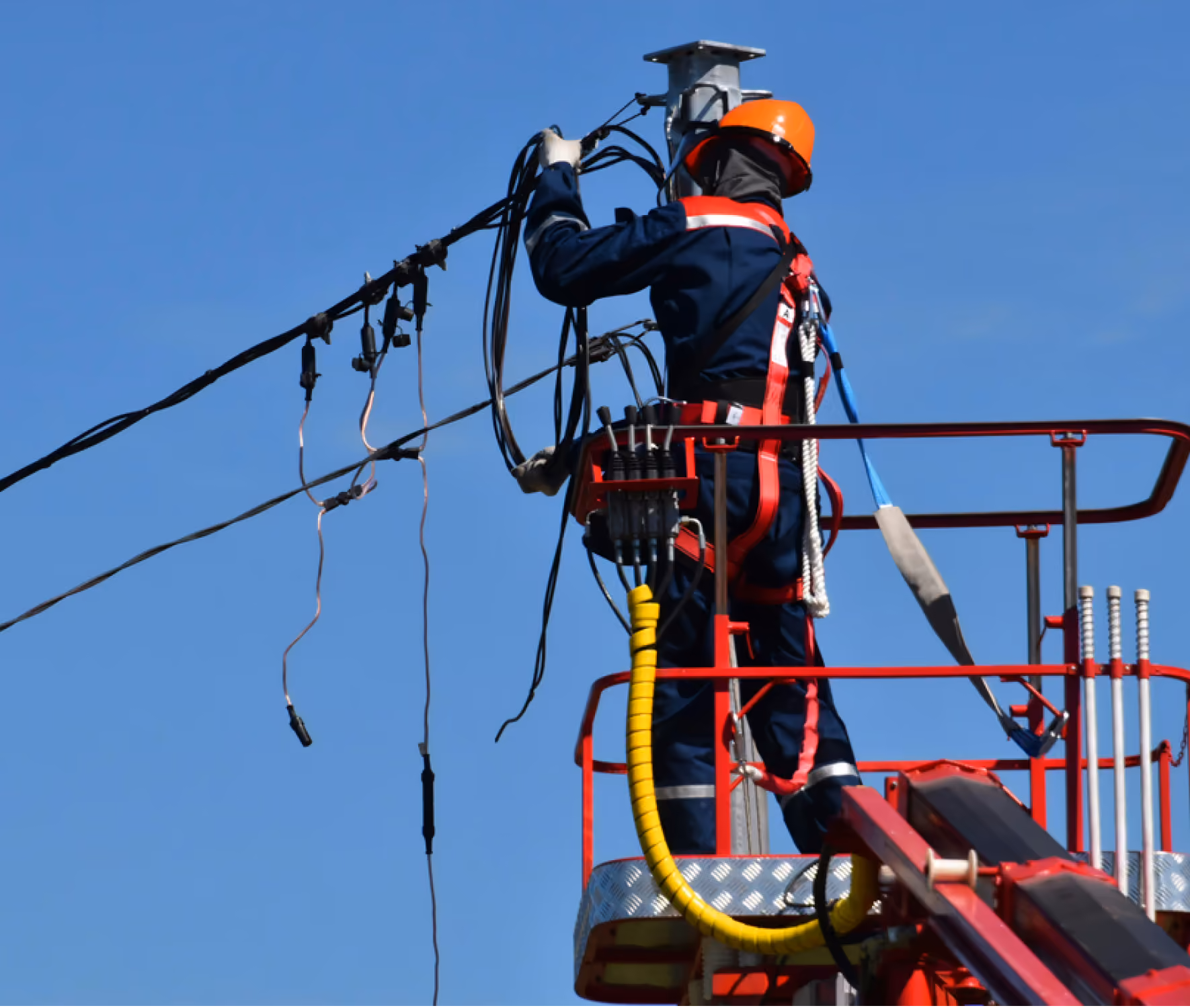 Electrician wearing safety gear working on power lines from a red elevated platform against blue sky.