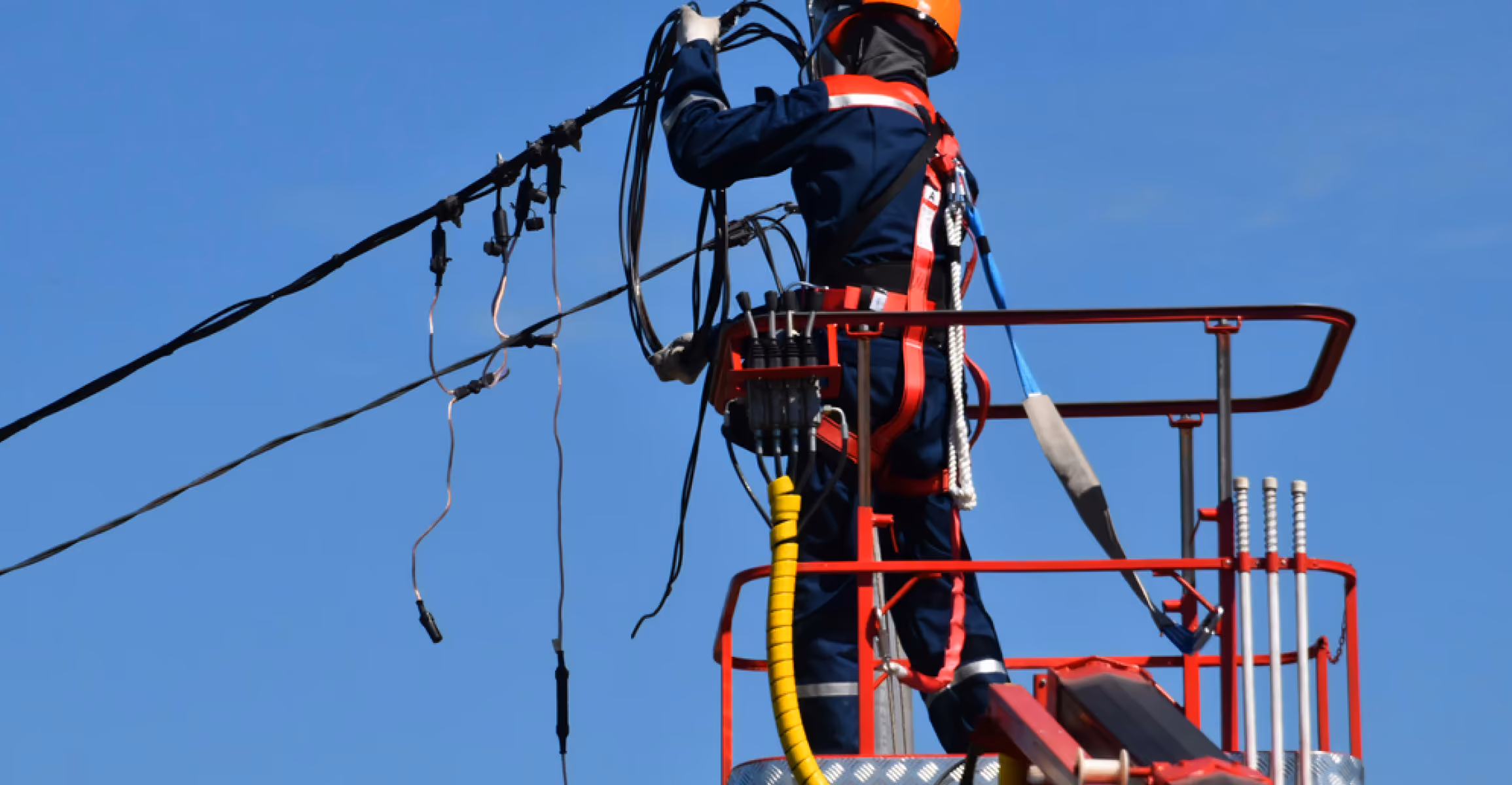 Worker in safety gear repairing electrical cables on an elevated platform against clear blue sky.