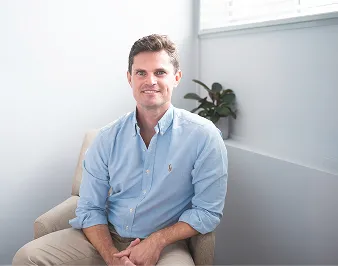 Smiling man in light blue shirt sitting on beige couch in a bright room with a potted plant.