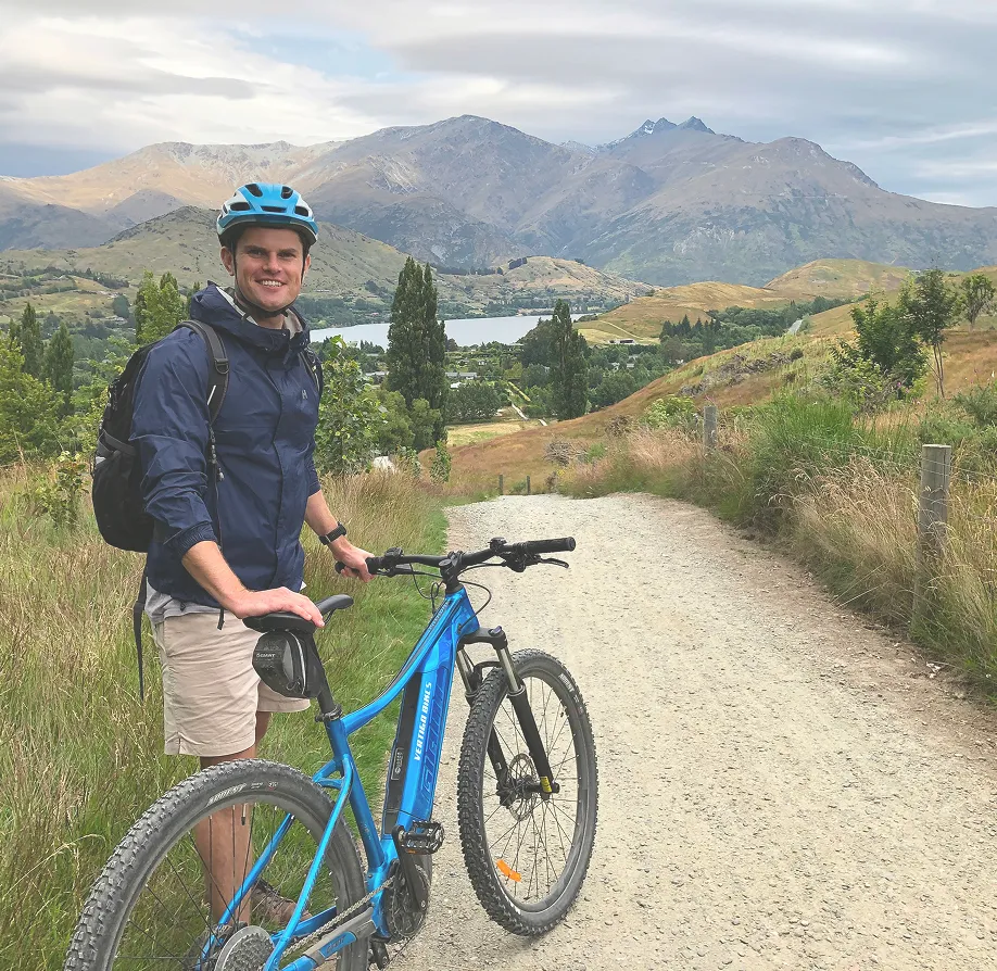 Smiling man in helmet standing with blue mountain bike on a gravel path with hills and lake behind.