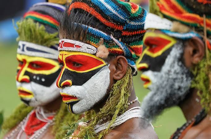 Three people with intricate red, black, and white facial paint and green moss decorations in colorful headdresses.