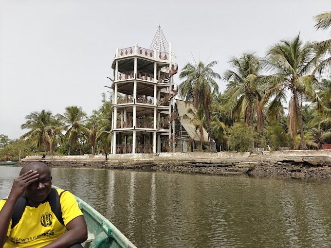 Person paddling a small boat on water near wooden houses with green roofs surrounded by trees.
