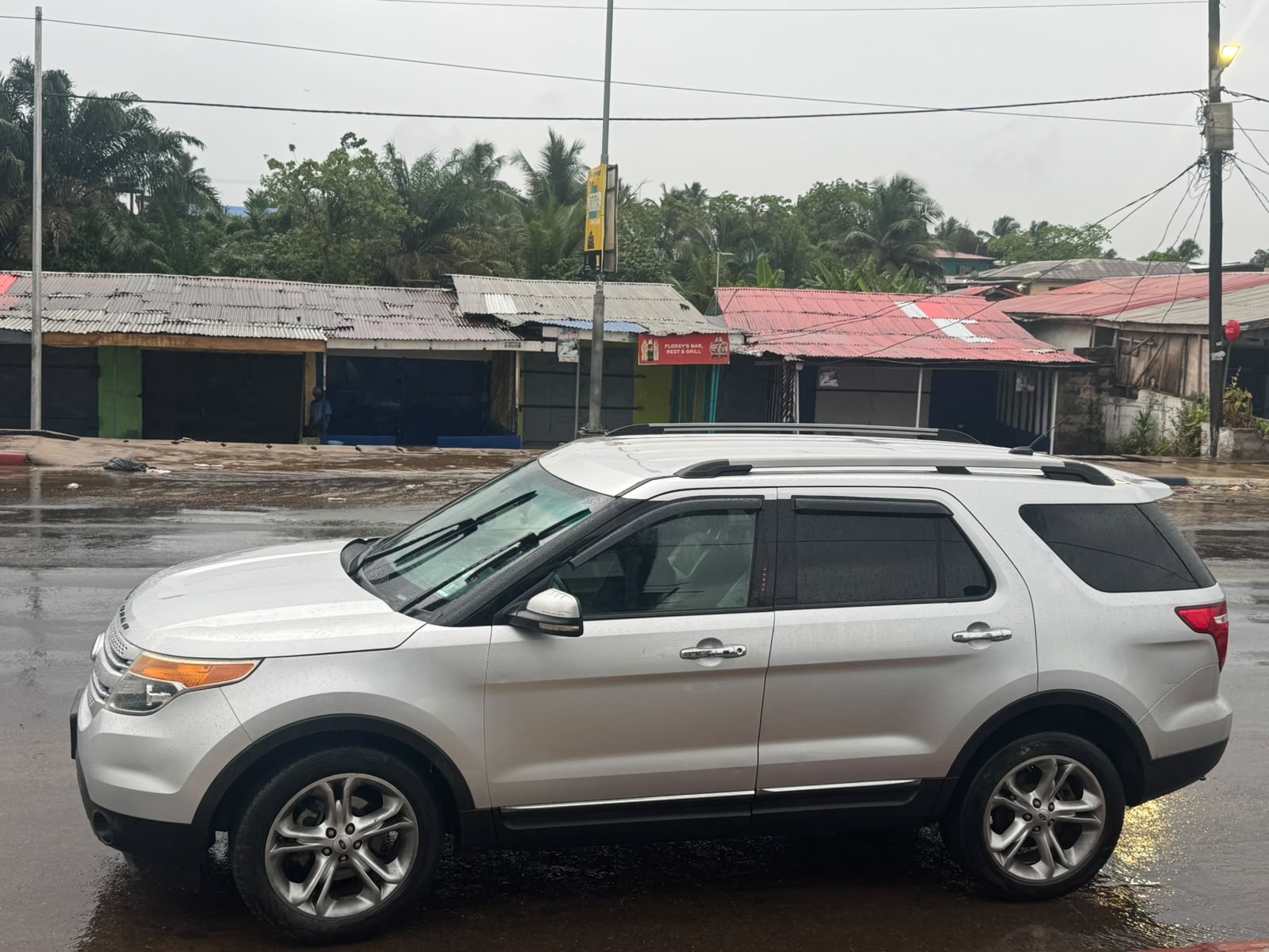 Silver SUV parked on a wet road in front of small buildings with corrugated metal roofs and greenery in the background.
