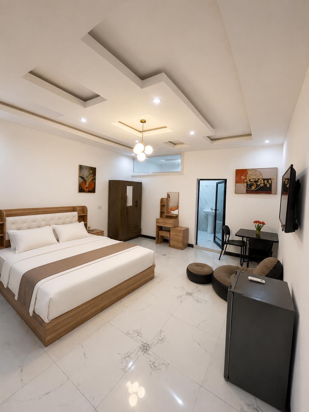 Modern bedroom with a large bed featuring white and beige bedding, a wooden wardrobe, a vanity desk with a mirror, a small dining table with two chairs and flowers, a wall-mounted TV, a fridge with remote on top, and a door leading to a bathroom with white marble tiles.