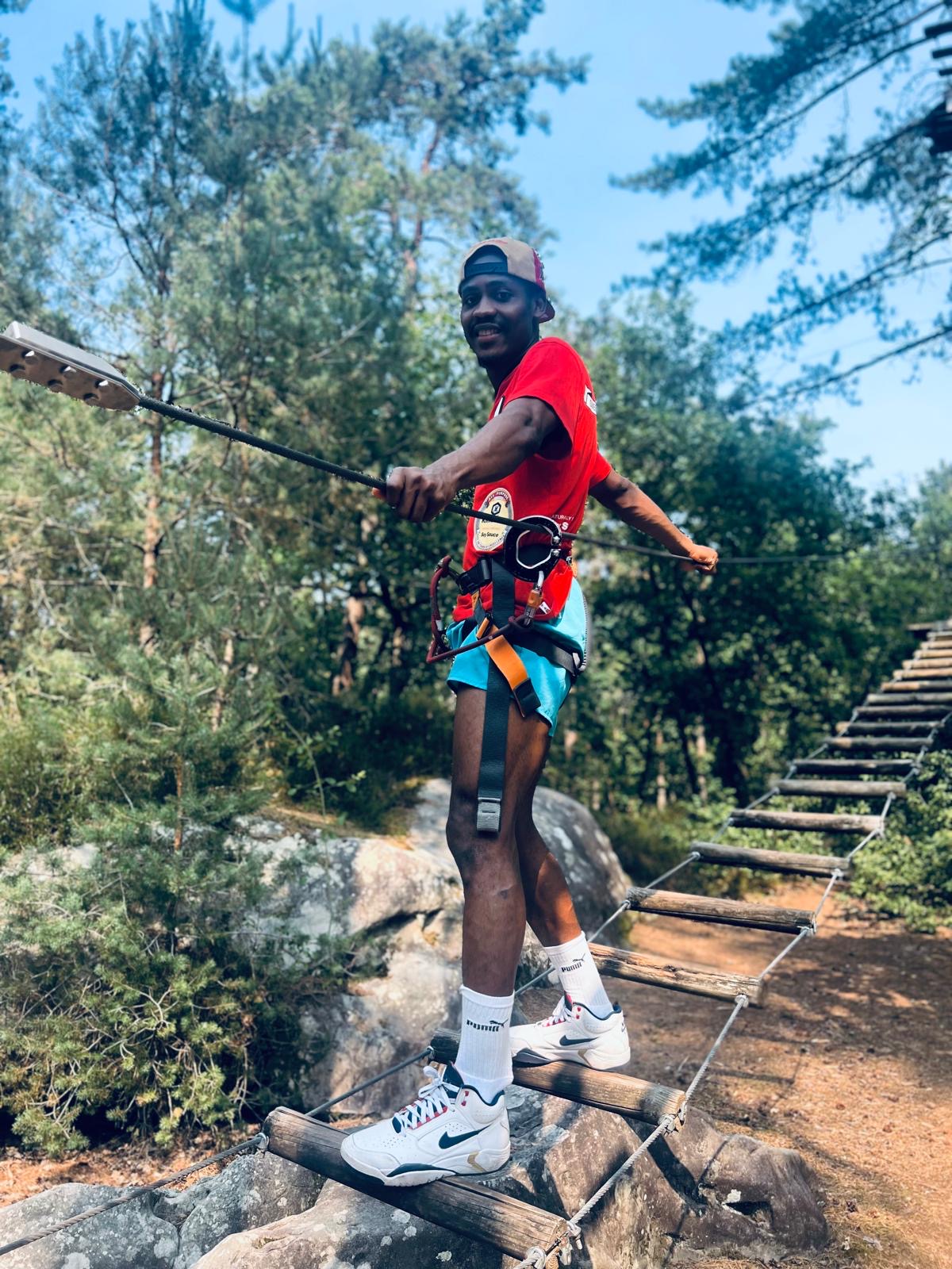 Man wearing a red shirt, blue shorts, and white Nike sneakers balancing on a wooden rope ladder suspended outdoors among trees, holding onto a safety harness attached to a cable.