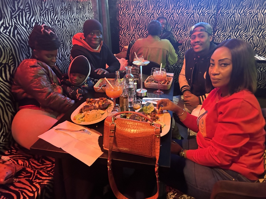 Group of six people sitting around a table at a restaurant with zebra print walls and upholstery, enjoying drinks and plates of food, including rice and grilled items, with an orange handbag placed on the table.
