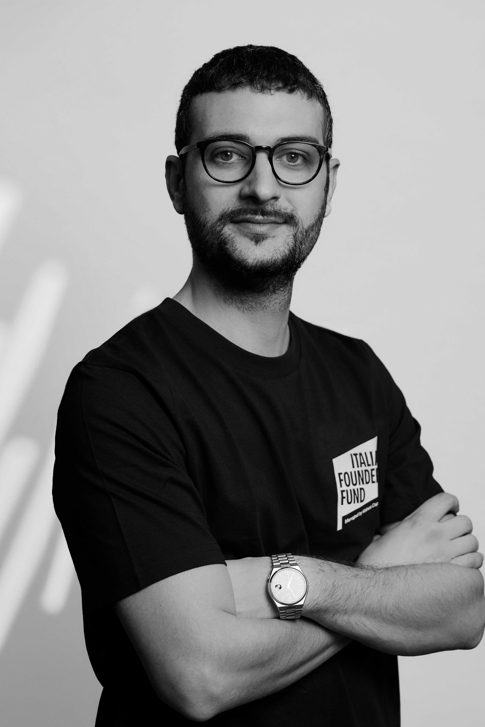 Man with short hair and glasses wearing a black shirt and watch, arms crossed, smiling slightly.