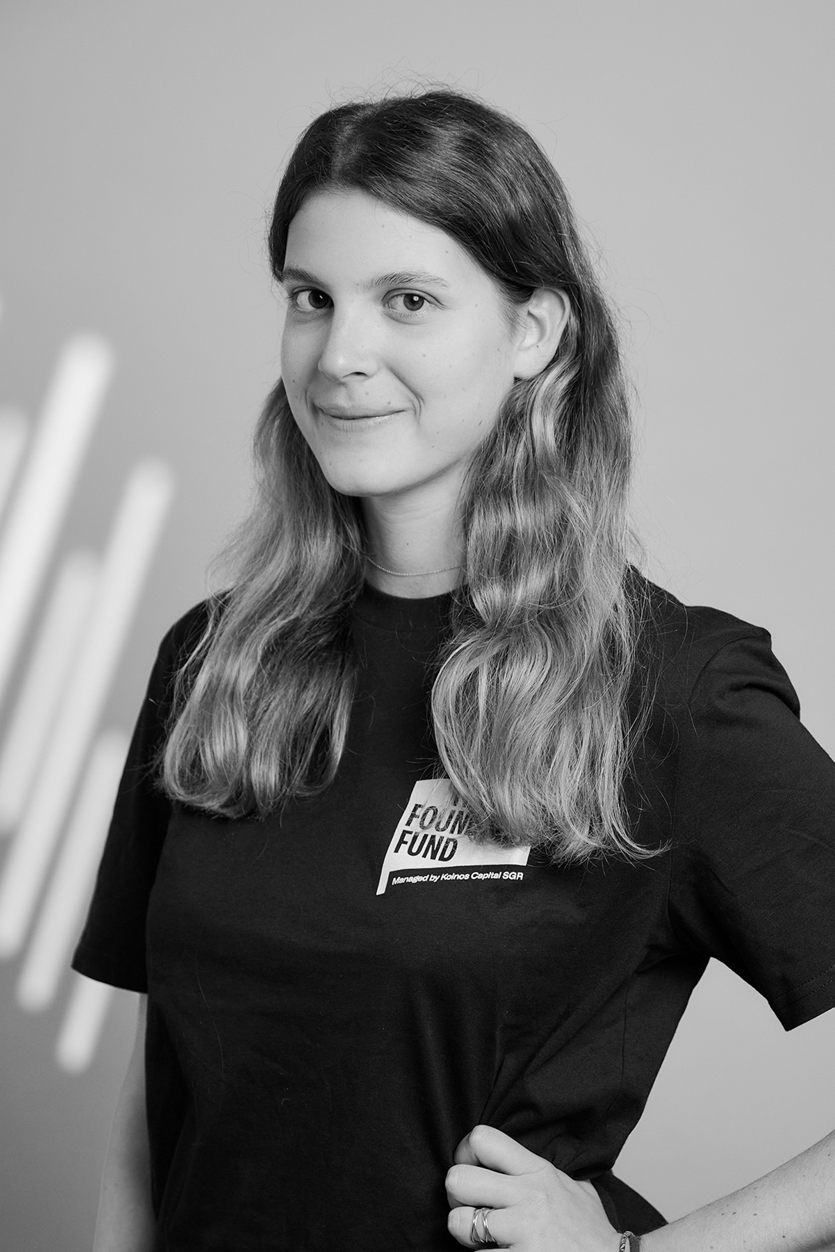 Young woman with wavy hair wearing a black shirt with 'Found Fund' text, posing with hand on hip.