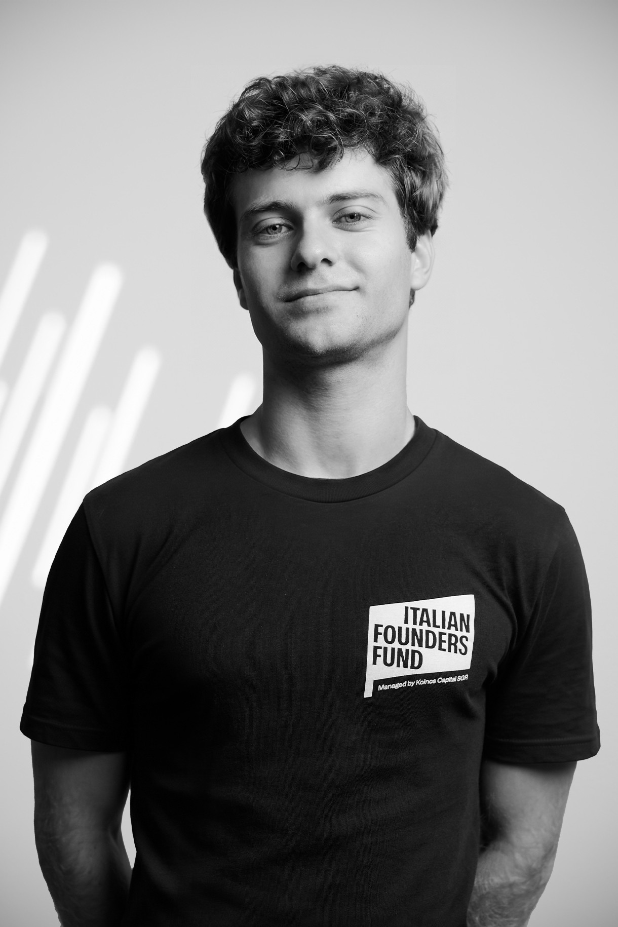 Young man with curly hair wearing a black Italian Founders Fund t-shirt, smiling slightly.