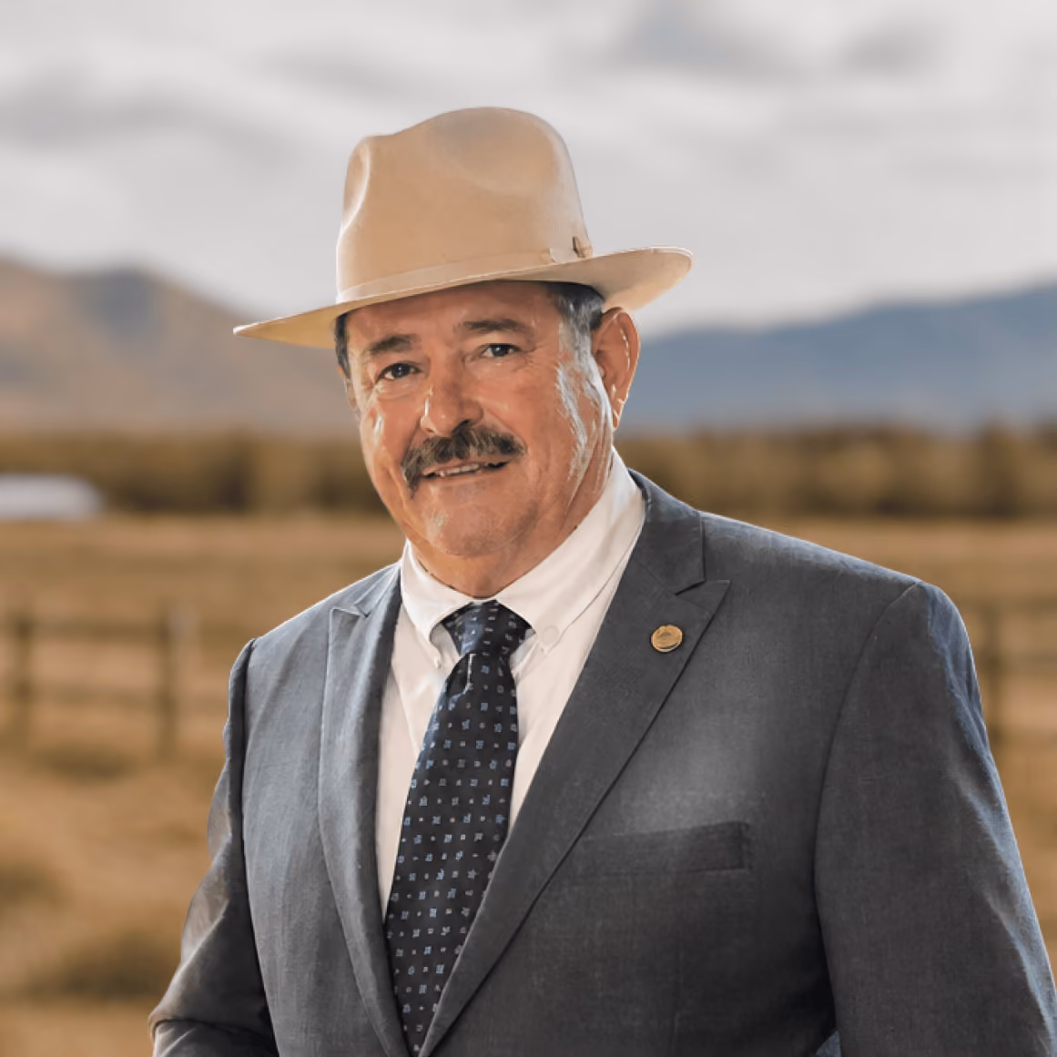 Michael Hogue in a suit and hat standing indoors with a bronze elk sculpture and mountain view behind him.