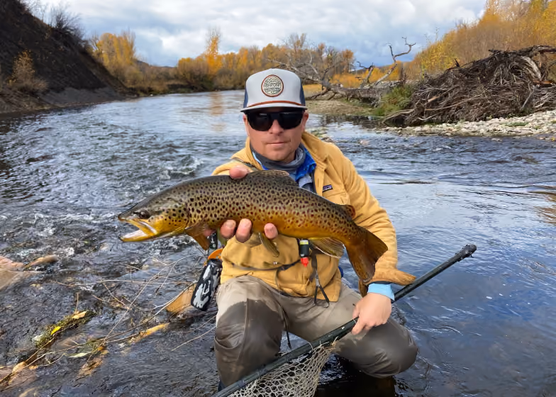 Man in sunglasses holding large brown trout while kneeling in shallow river with fall trees in background.