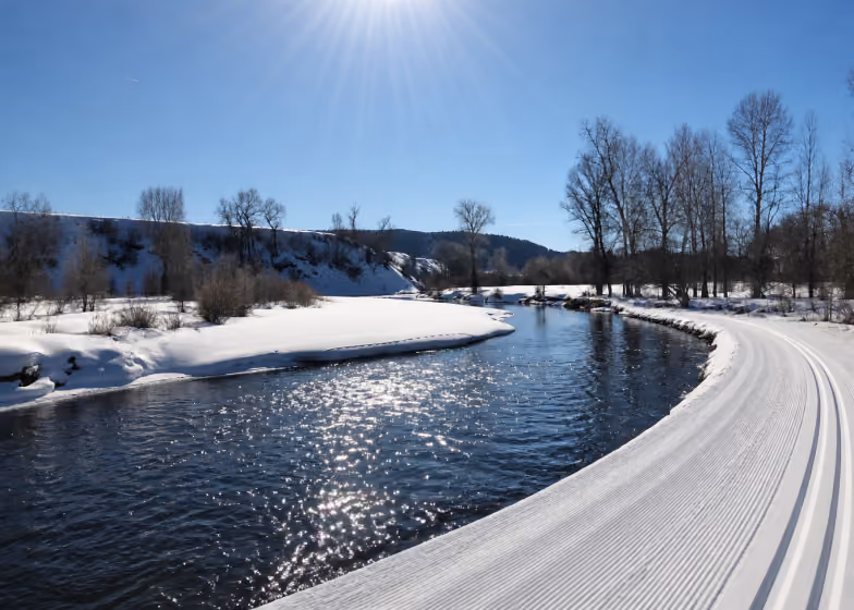 Sunlit river with snow-covered banks and groomed ski trails curving alongside in a clear winter scene.