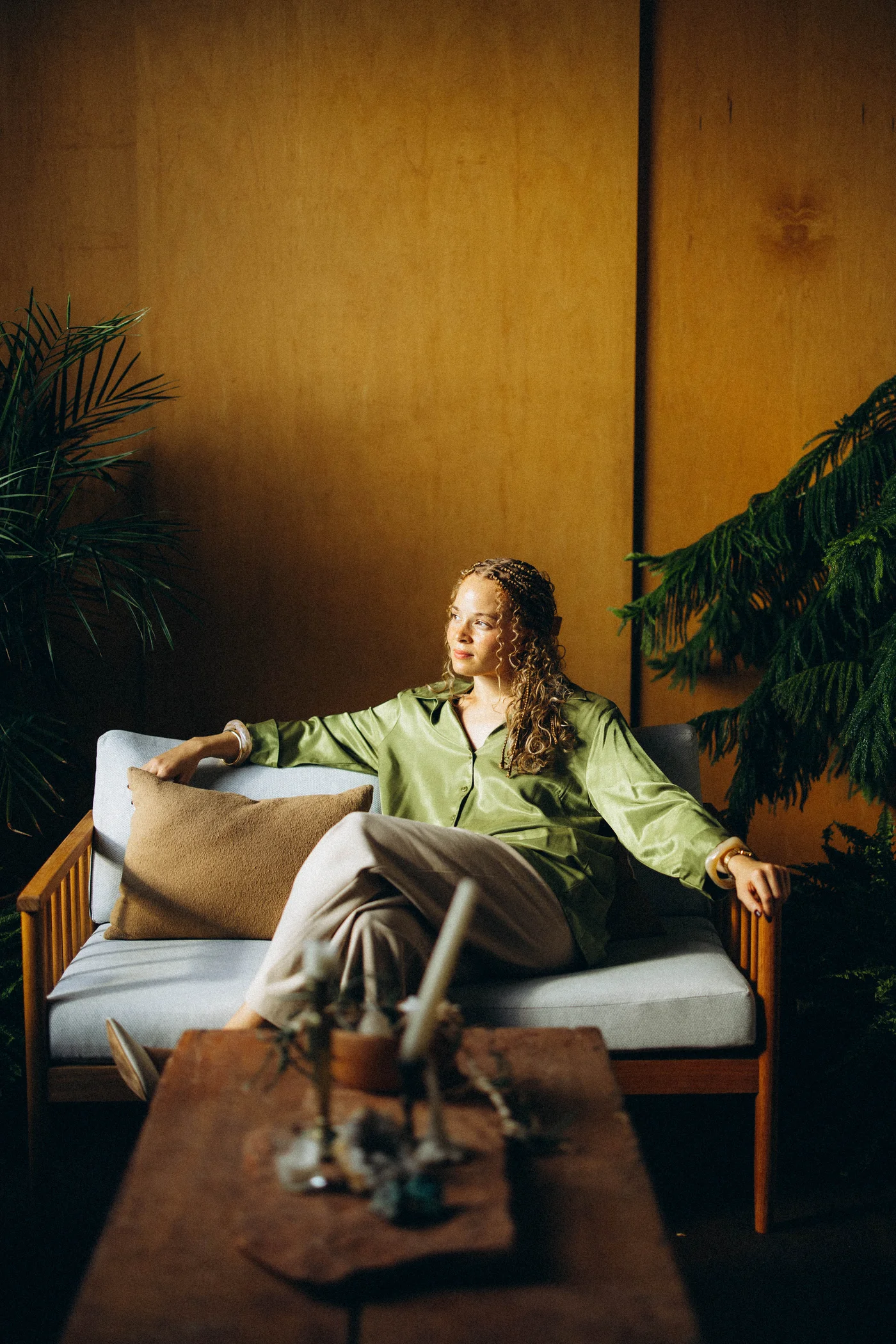Amber Williamson, founder of Anomaly, reclined on a wood-frame sofa in a green satin shirt, with tropical plants and warm golden wall behind her