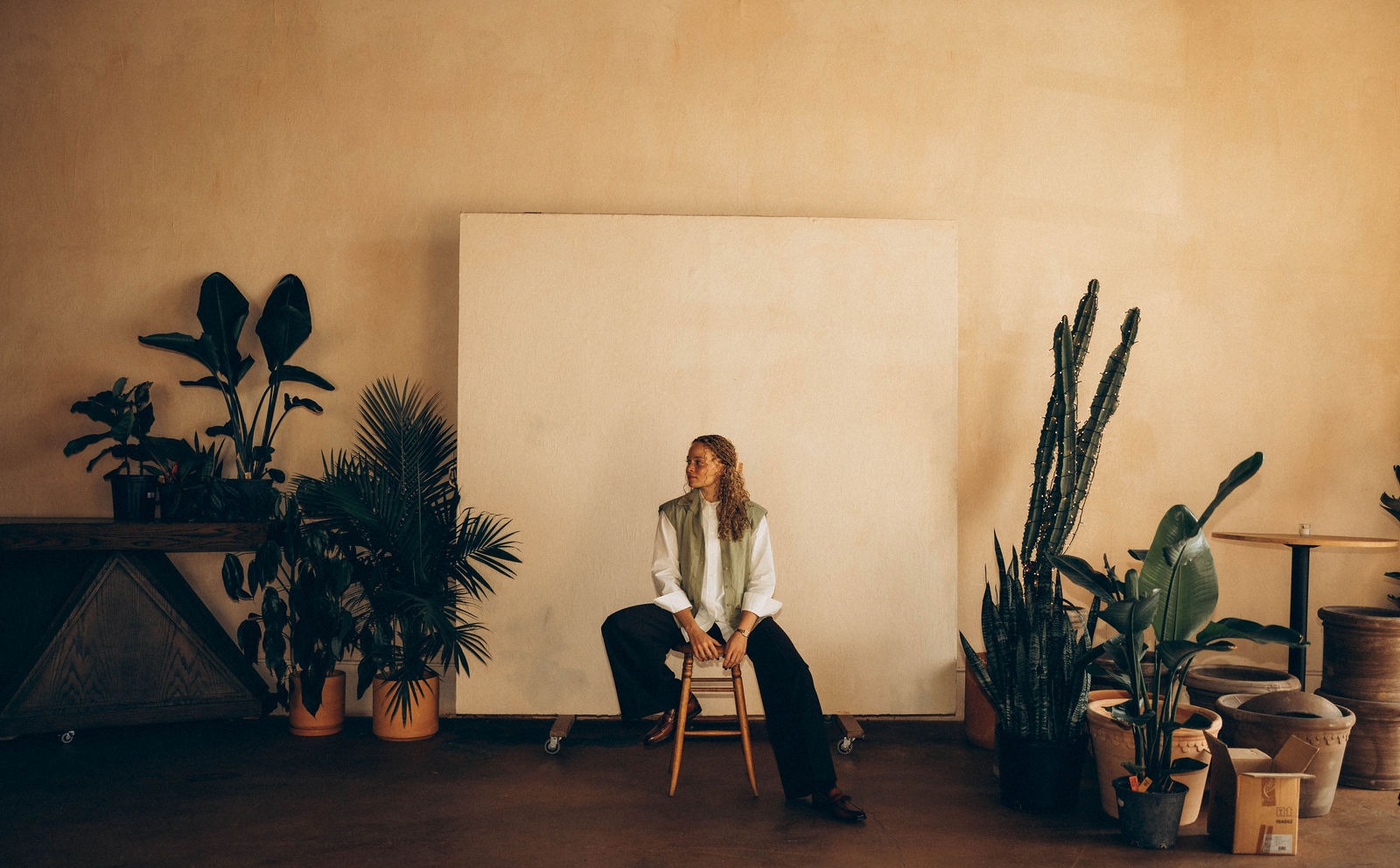 Amber Williamson, founder of Anomaly, seated on wooden stool surrounded by lush indoor plants in a warm studio setting