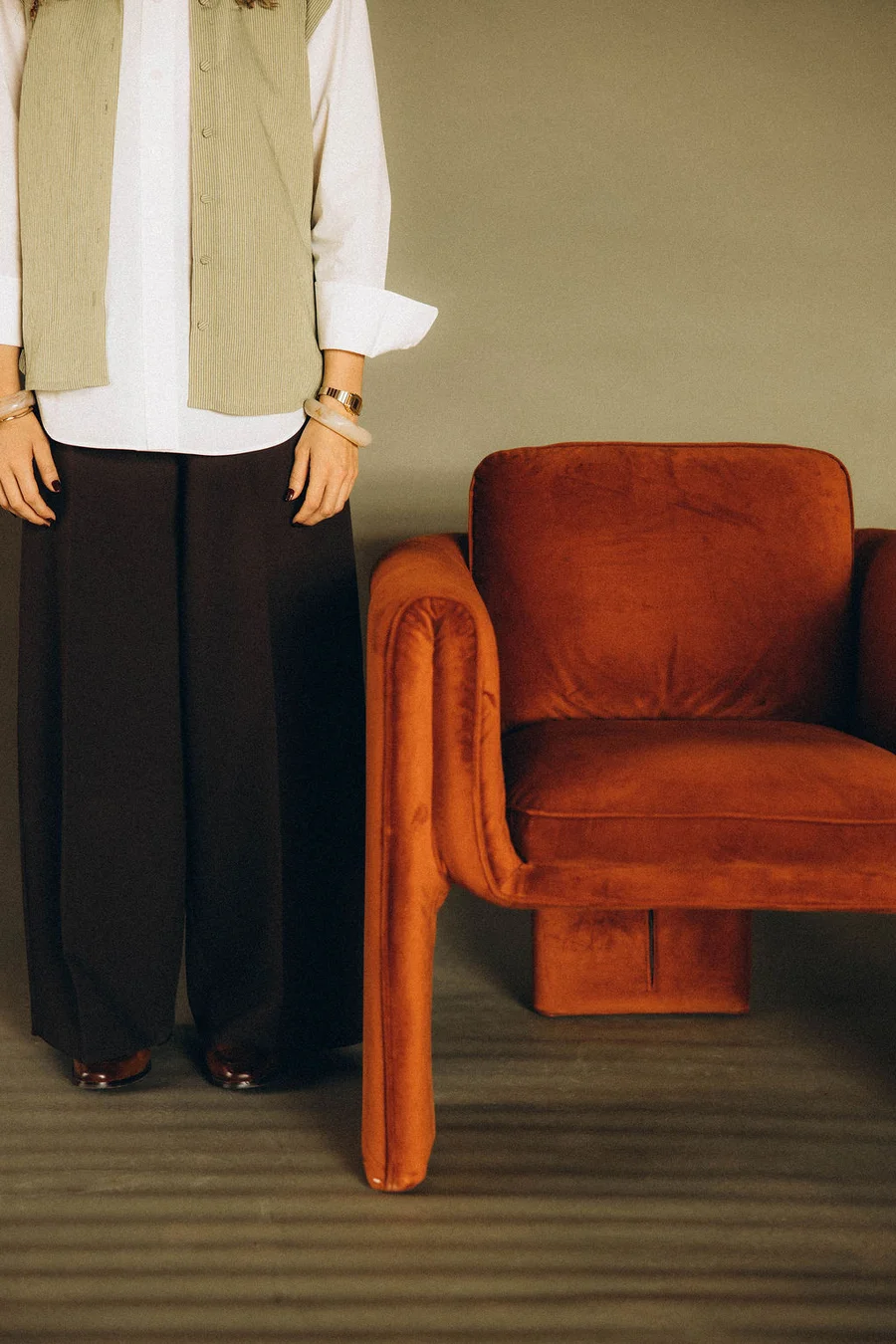 Close-up detail of Anomaly founder standing beside an orange velvet armchair, wearing a striped vest and wide-leg trousers with gold bracelets