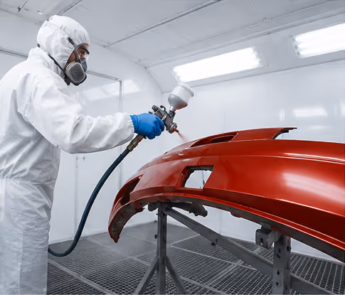 Person in white protective suit and mask spray-painting a red car bumper inside a paint booth.