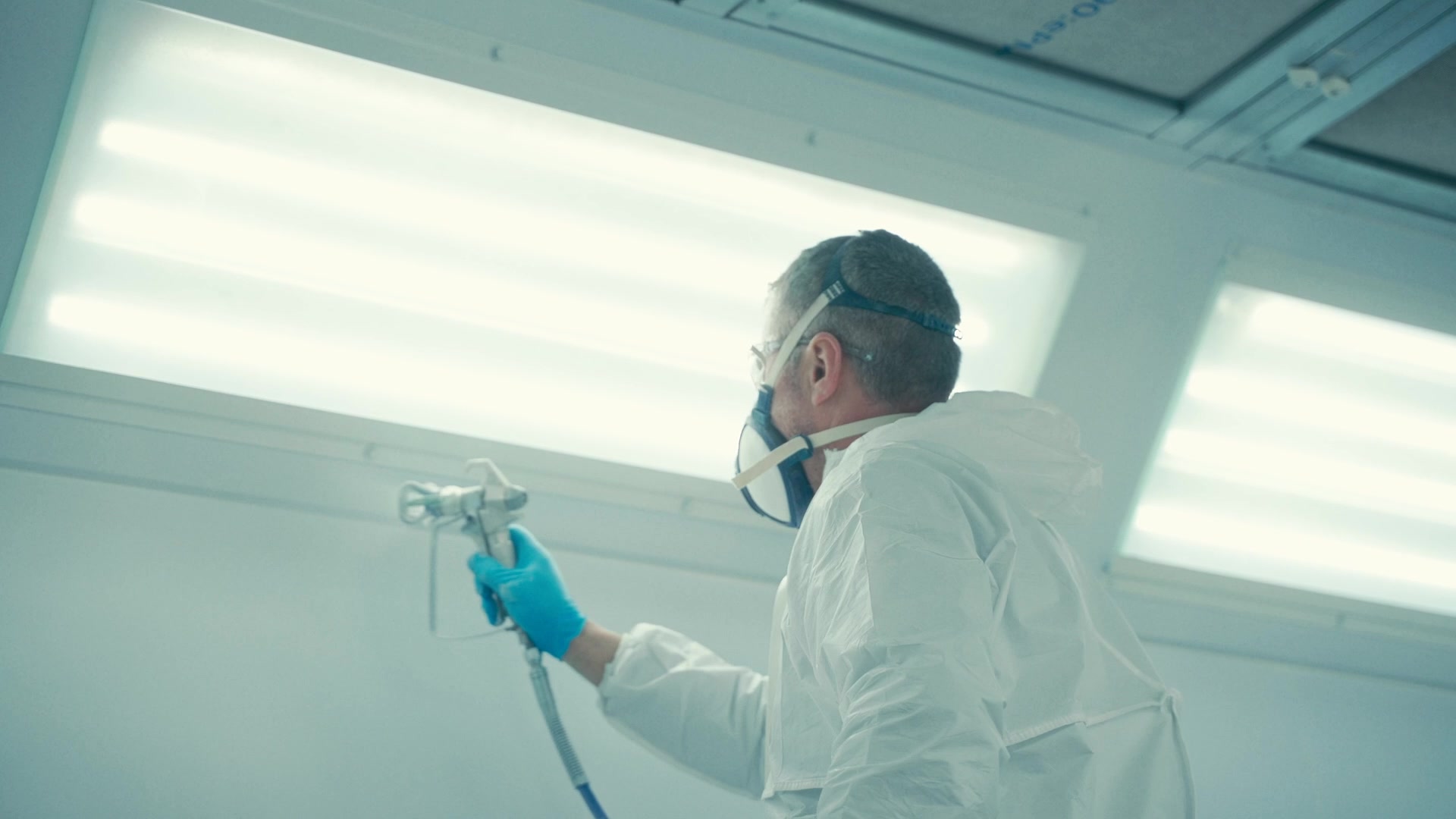 Man wearing protective white suit, blue gloves, and face mask using a spray gun to paint or coat an indoor surface.