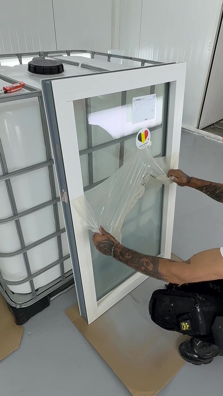 Person peeling protective plastic off a white-framed window standing vertically on the floor near a large white tank container with metal framework.