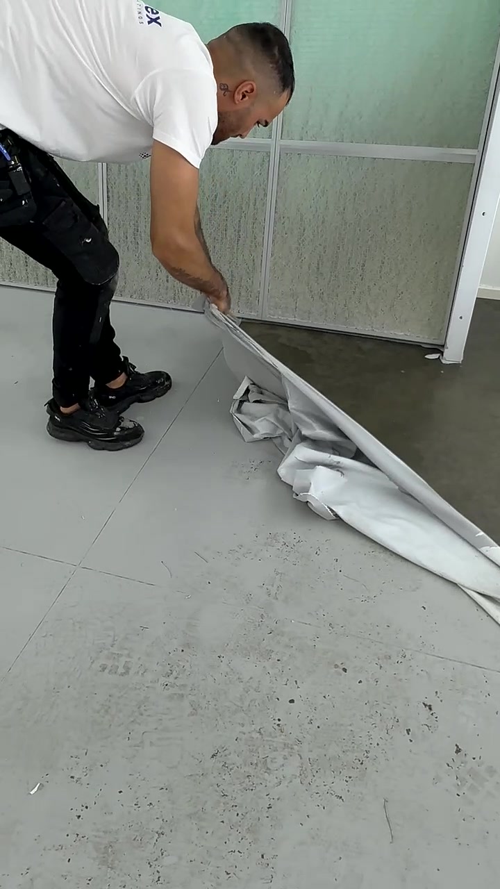 Man in black pants and black shoes lifting and peeling back a large sheet of gray flooring from a gray tiled surface.
