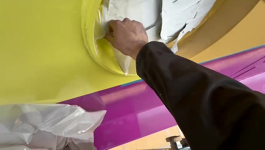 A person in a black jacket peeling white protective plastic film off a yellow and purple surface.