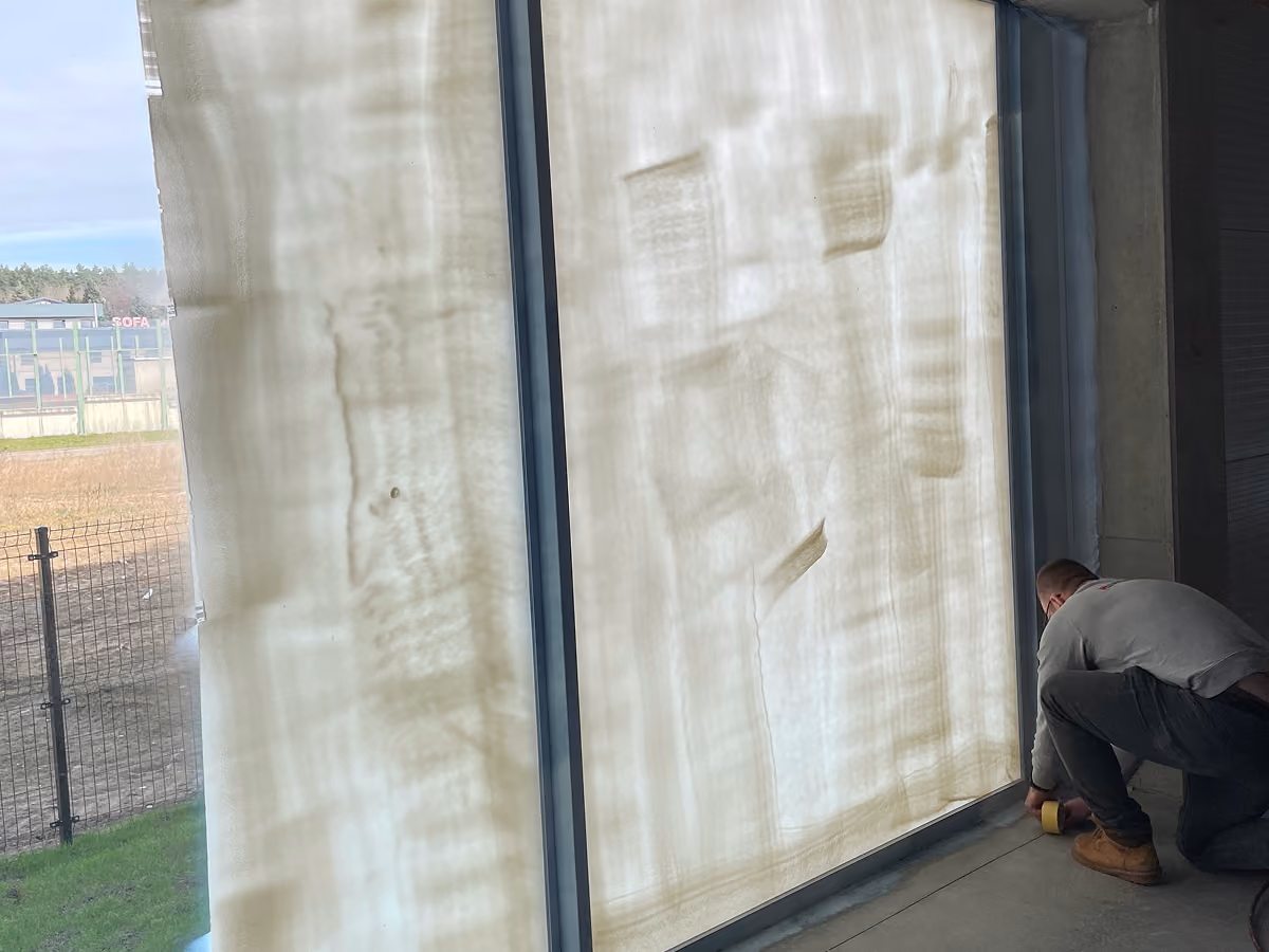 Man kneeling on floor taping the bottom edge of large glass window covered with peelable coating inside building.