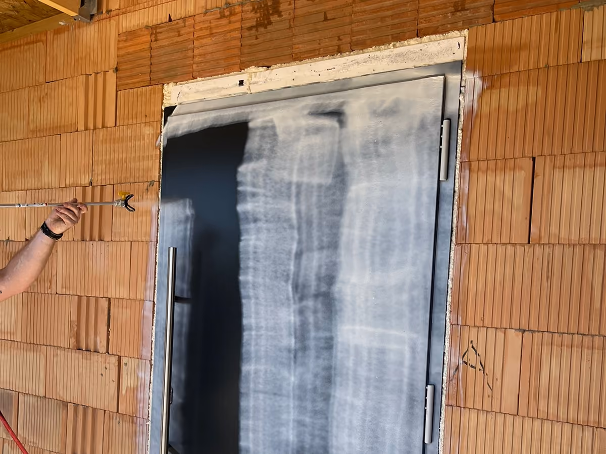 Person spraying white peelable coating over a black metal door frame on a brick wall.