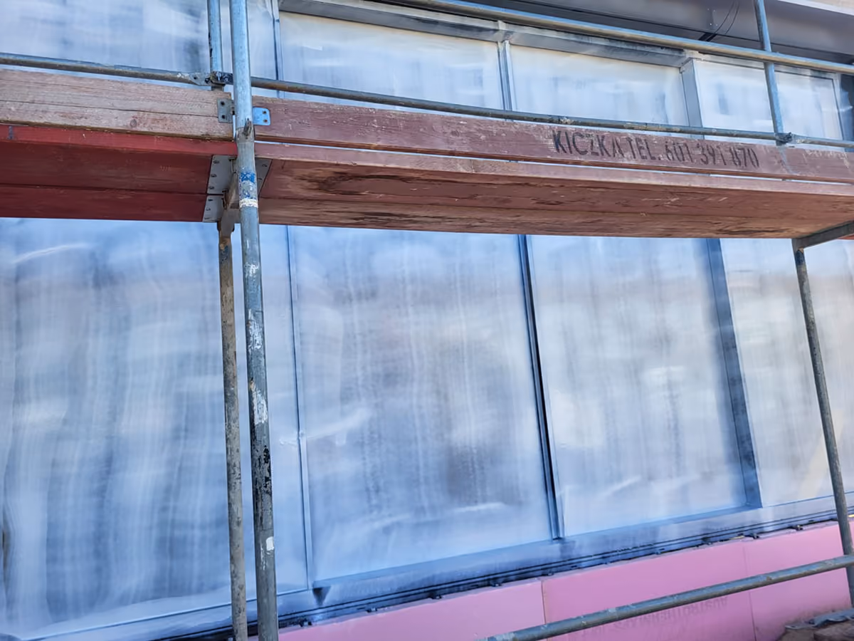Metal scaffolding with a wooden plank set up against a wall with windows covered with peelable coating.