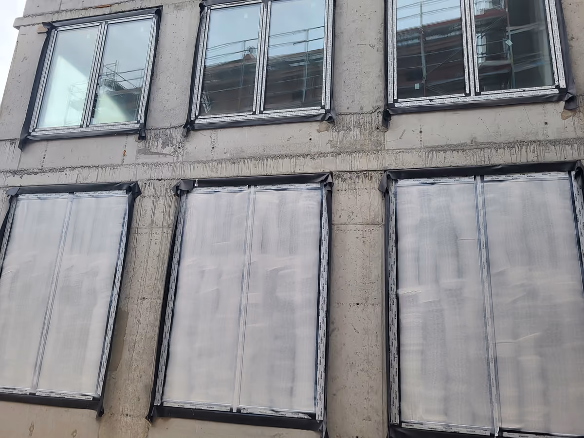 Concrete building facade with six windows in two rows; three upper windows show reflections and scaffolding, three lower windows are covered with protective plastic sheeting.