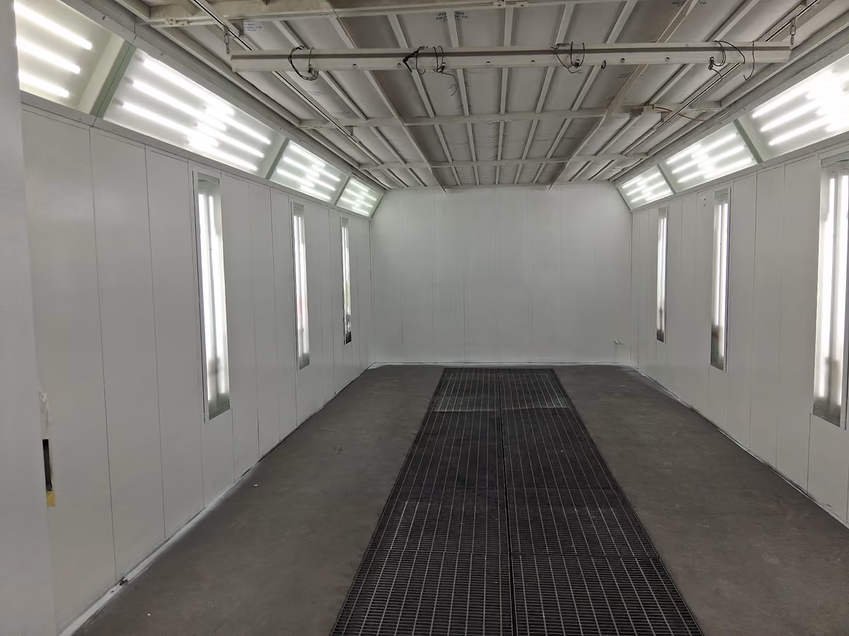 Empty industrial paint spray booth with bright overhead and side panel lights and grated floor vents.