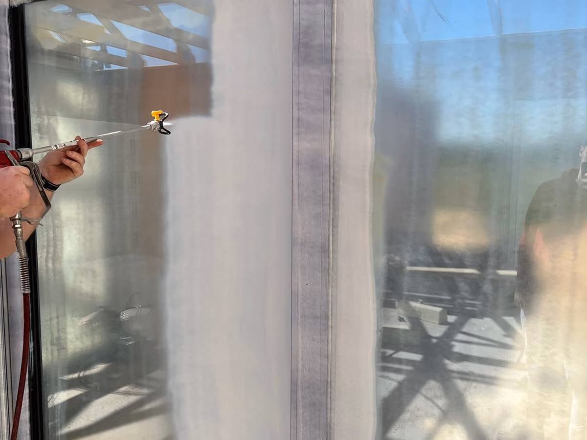 Person using a spray gun to apply protective coating on a large glass window.