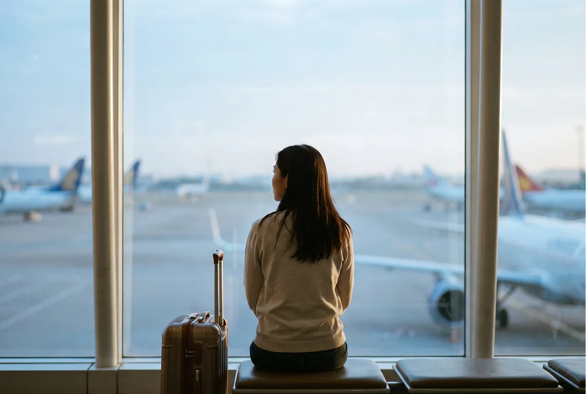 공항 터미널 창가에 캐리어를 두고 활주로를 바라보며 앉아 있는 여행자의 뒷모습