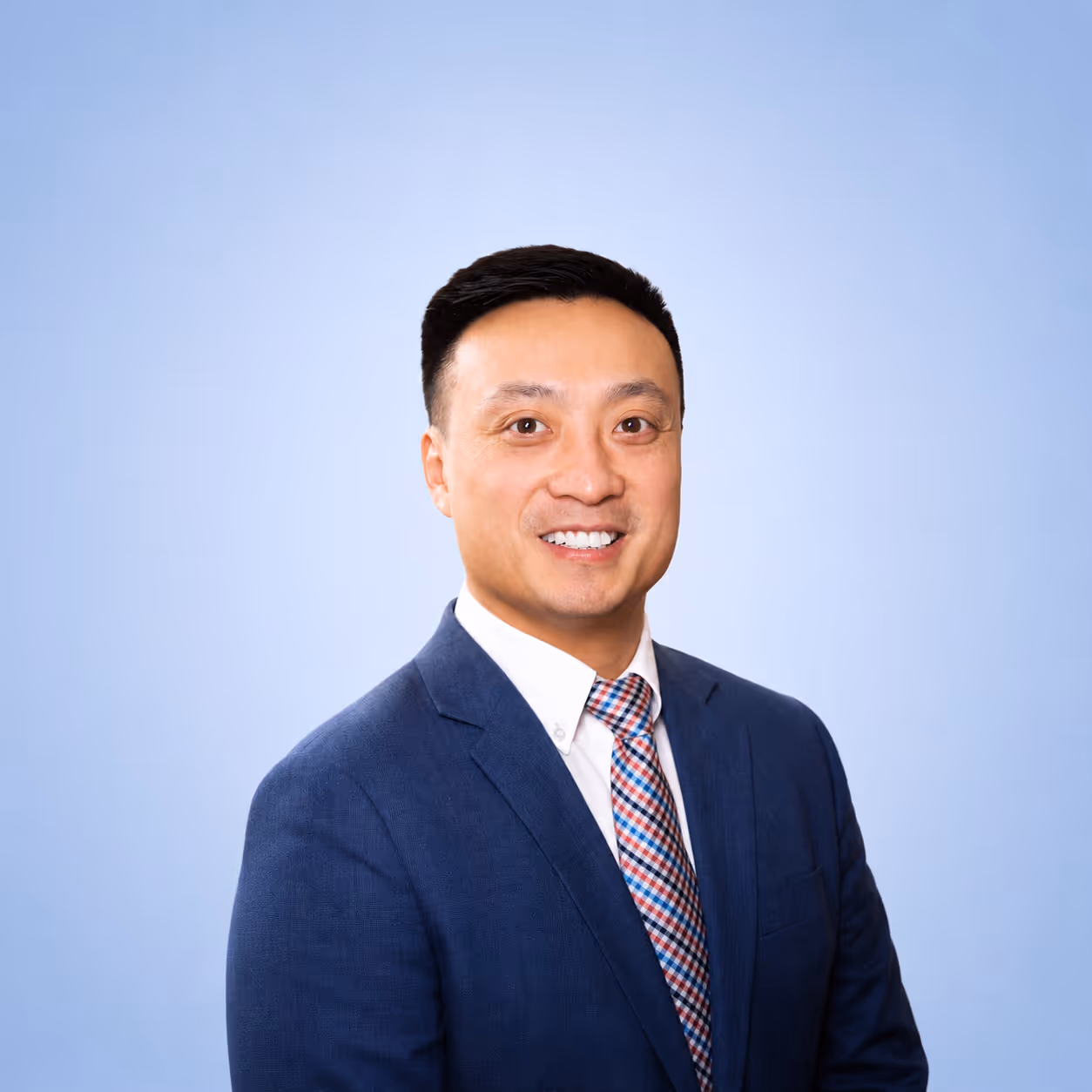 Smiling man in a blue suit jacket, white shirt, and checked tie against a light blue background.