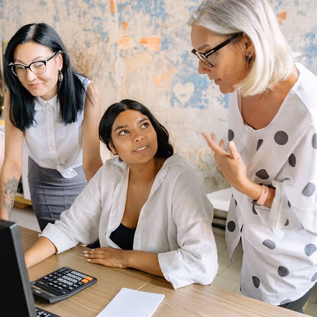 Three women in conversation at a desk with a calculator and computer monitor.