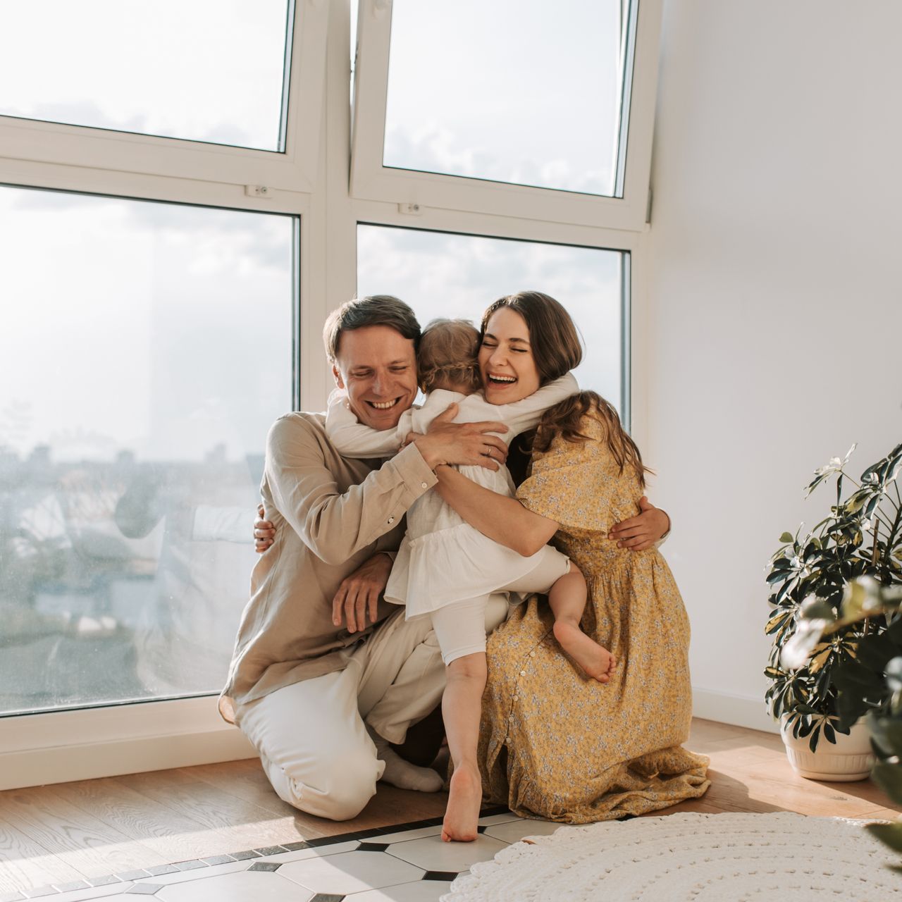 A smiling couple hugging their young child near a large window in a bright room.