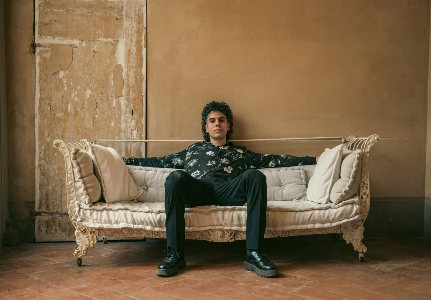 Editorial portrait of a man in black seated on a vintage sofa against a plaster wall, by GROOVE Media