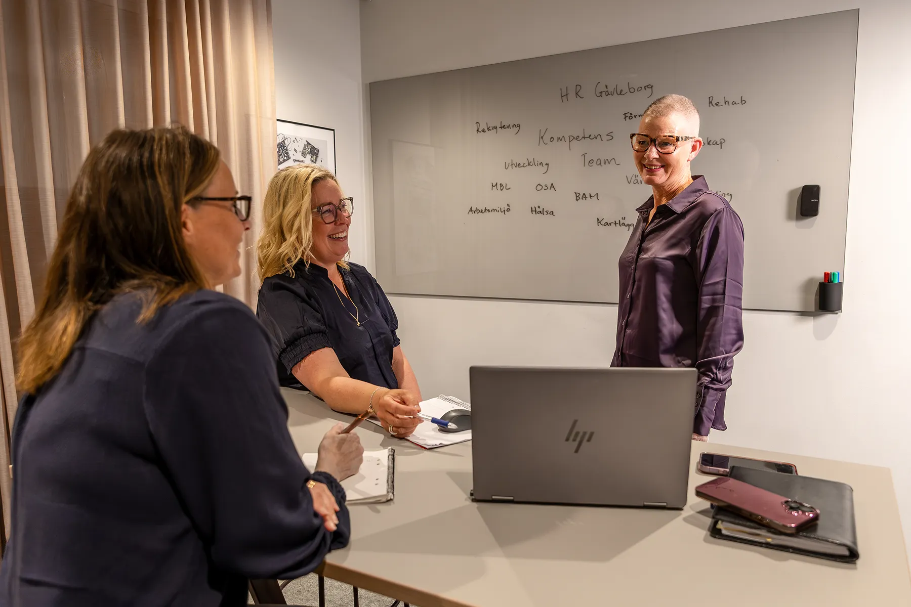 Tre kvinnor från HR Gävleborg i mötesrum med laptop och whiteboard med handskrivna ord bakom dem.