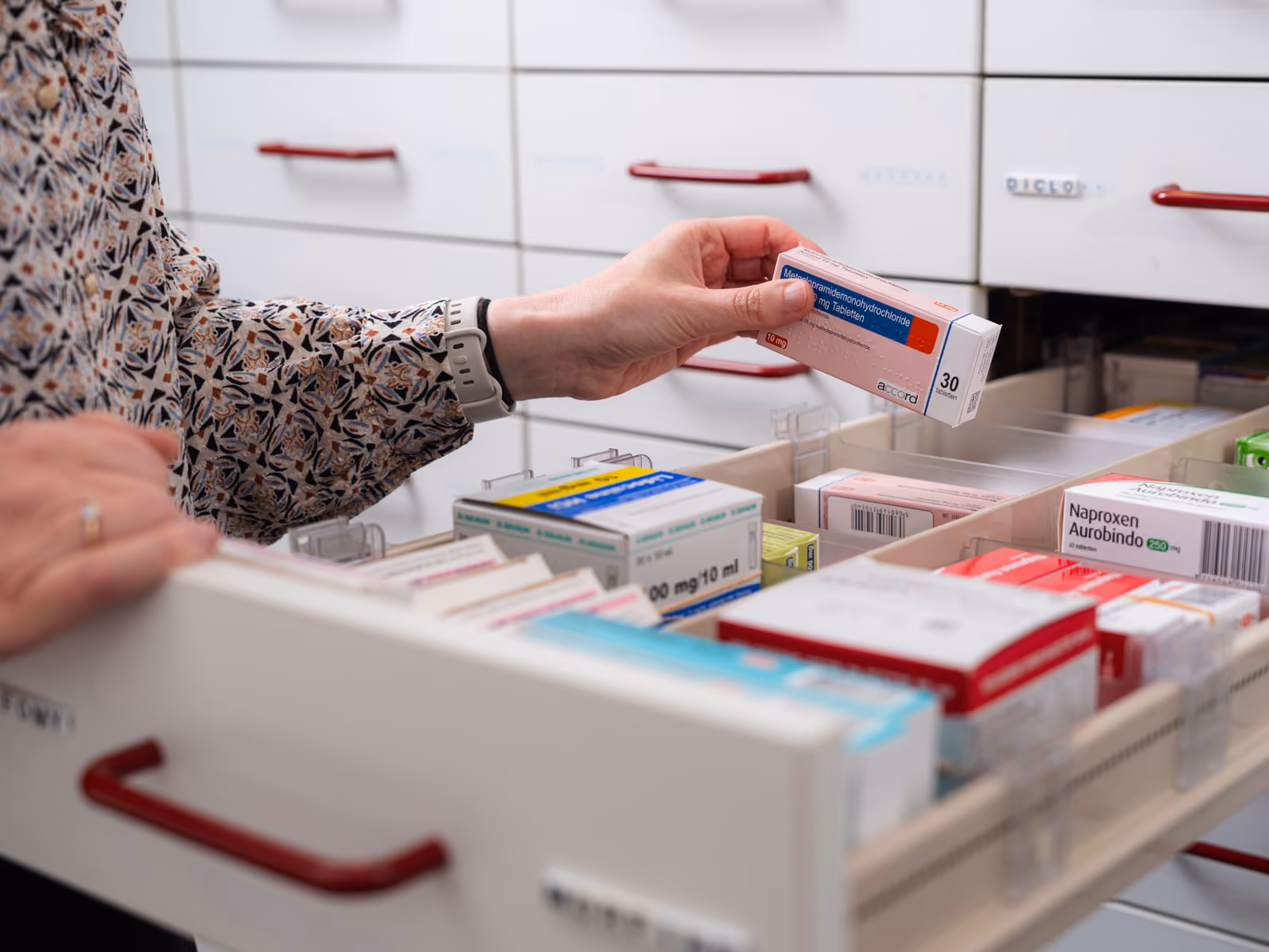 Iemand pakt een geneesmiddelenverpakking uit een apotheekkastlade met diverse medicijnendozen.