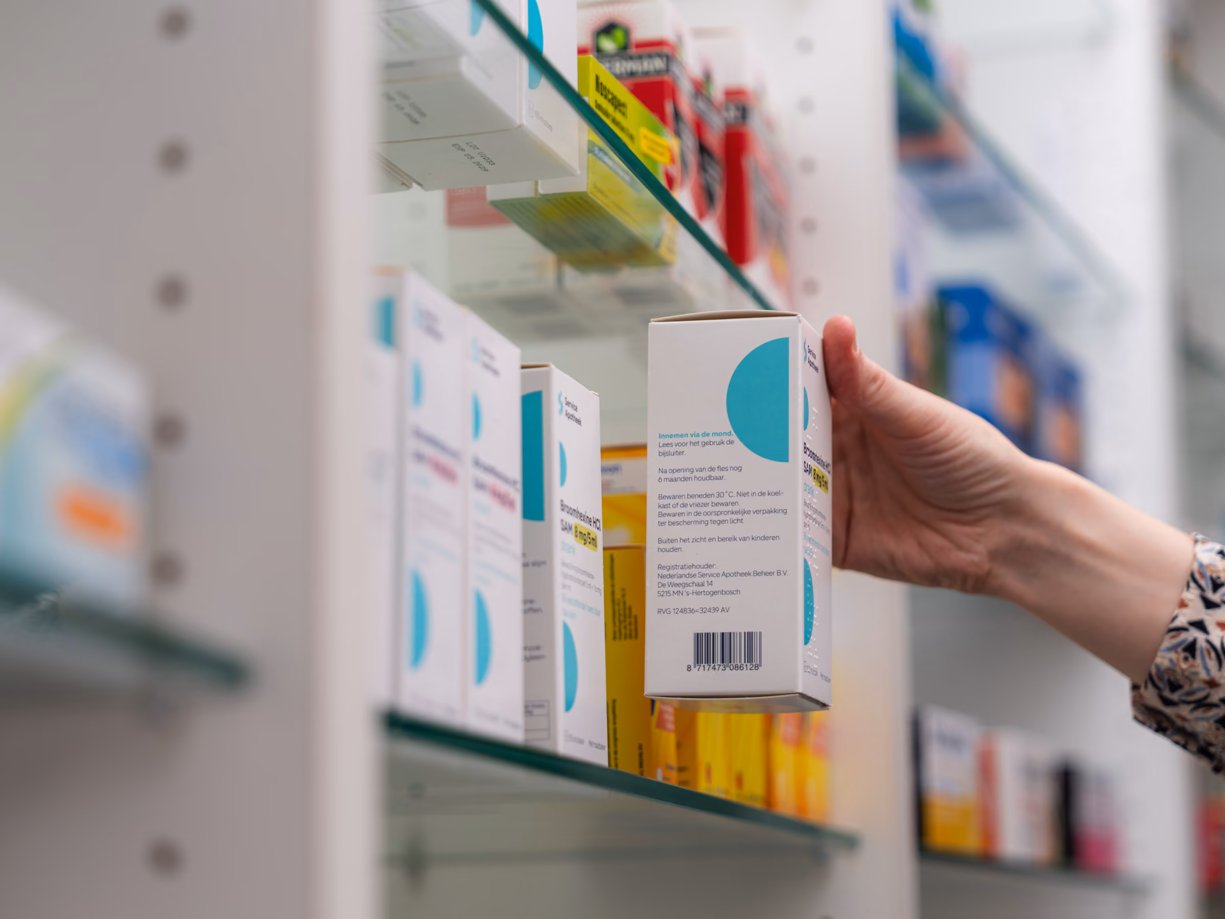 Hand pakt een doos medicatie van een glazen plank in een apotheek, omringd door andere medicijnendozen.
