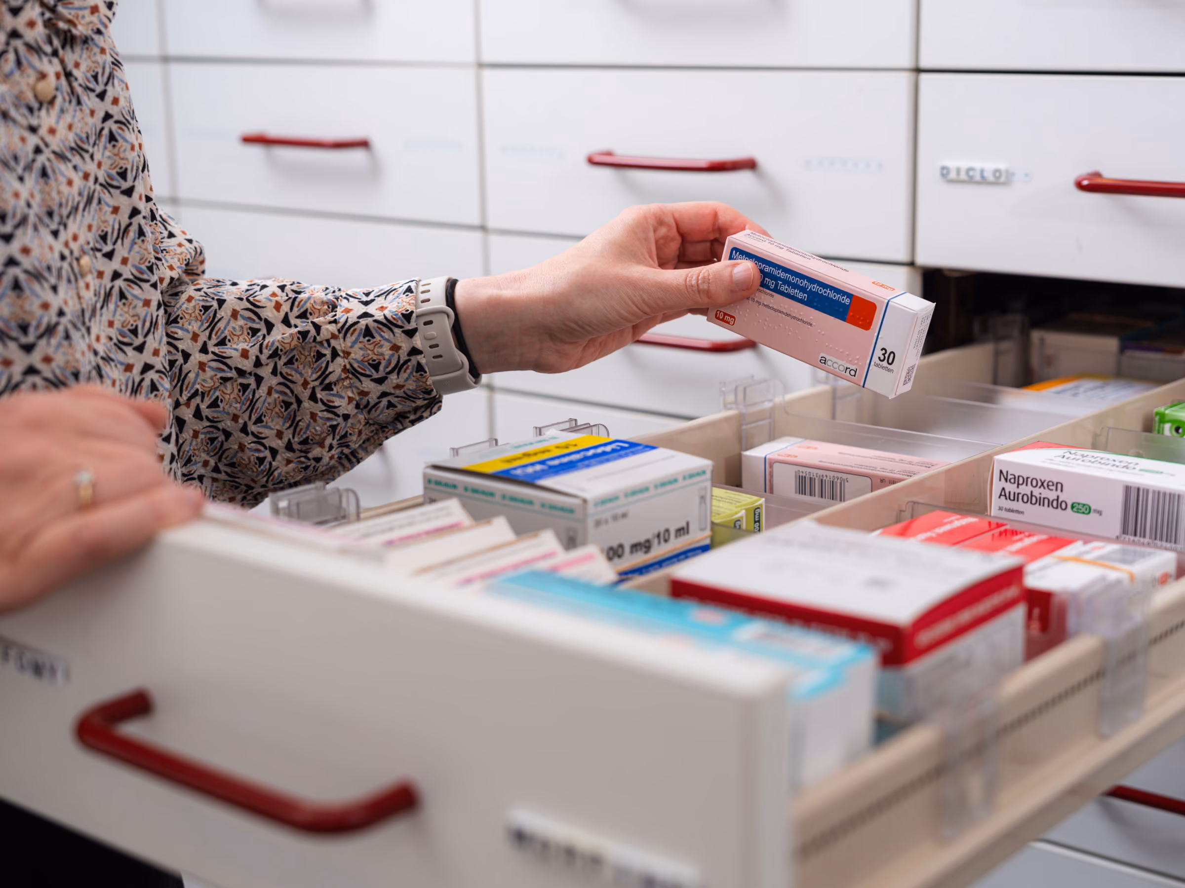 Een persoon pakt een doosje medicatie uit een kastlade gevuld met verschillende medicijnen in een apotheekomgeving.