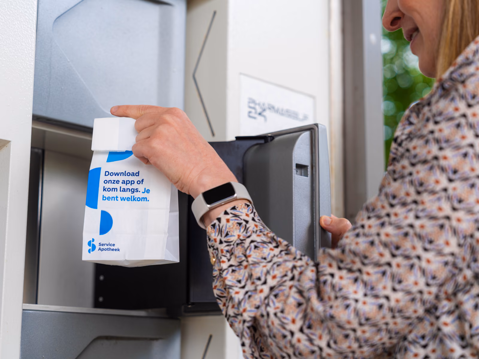 Vrouw die een wit papieren tasje met blauwe tekst uithaalt uit een pakketautomaat. Op het tasje staat: 'Download onze app of kom langs. Je bent welkom.'