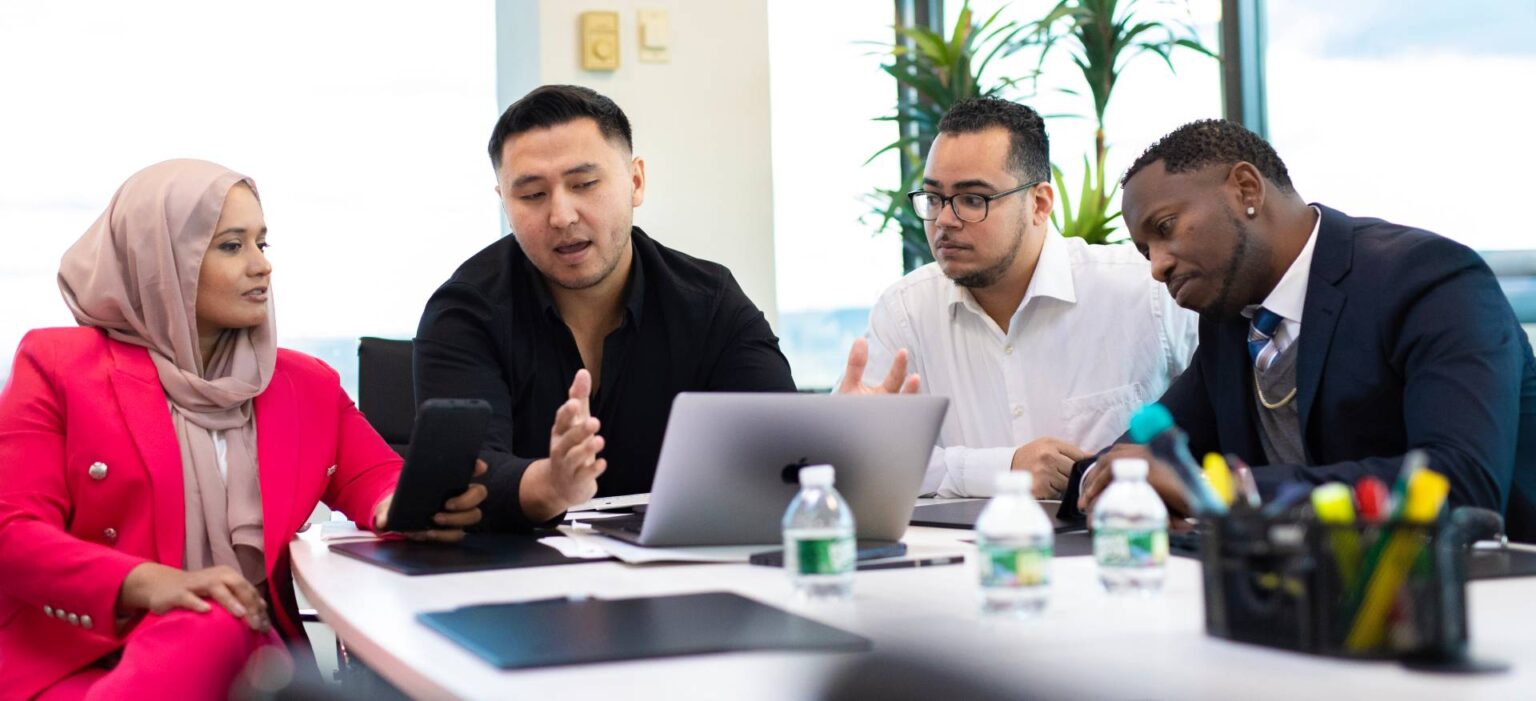 Diverse group in business attire discussing work with laptop and smartphone at a conference table.