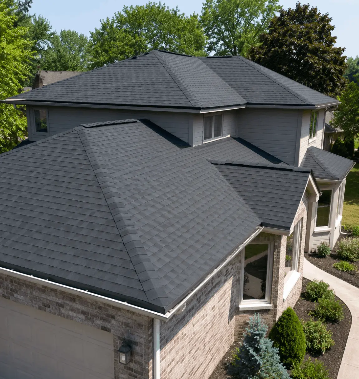 Dark grey shingle roof on a two-story stone home with hipped rooflines and landscaped front beds.