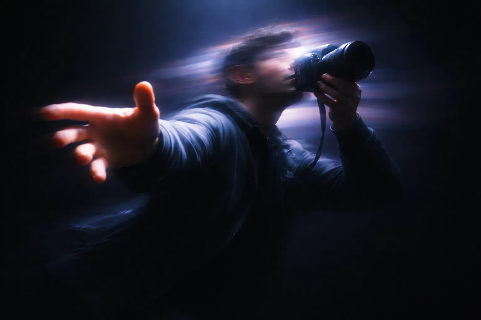 Blurred image of a person holding a camera and extending their hand in a dark setting.