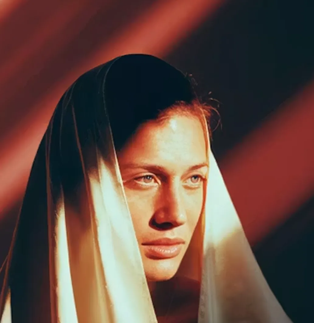 Close-up of a woman with blue eyes wearing a white headscarf against warm, shadowed background.